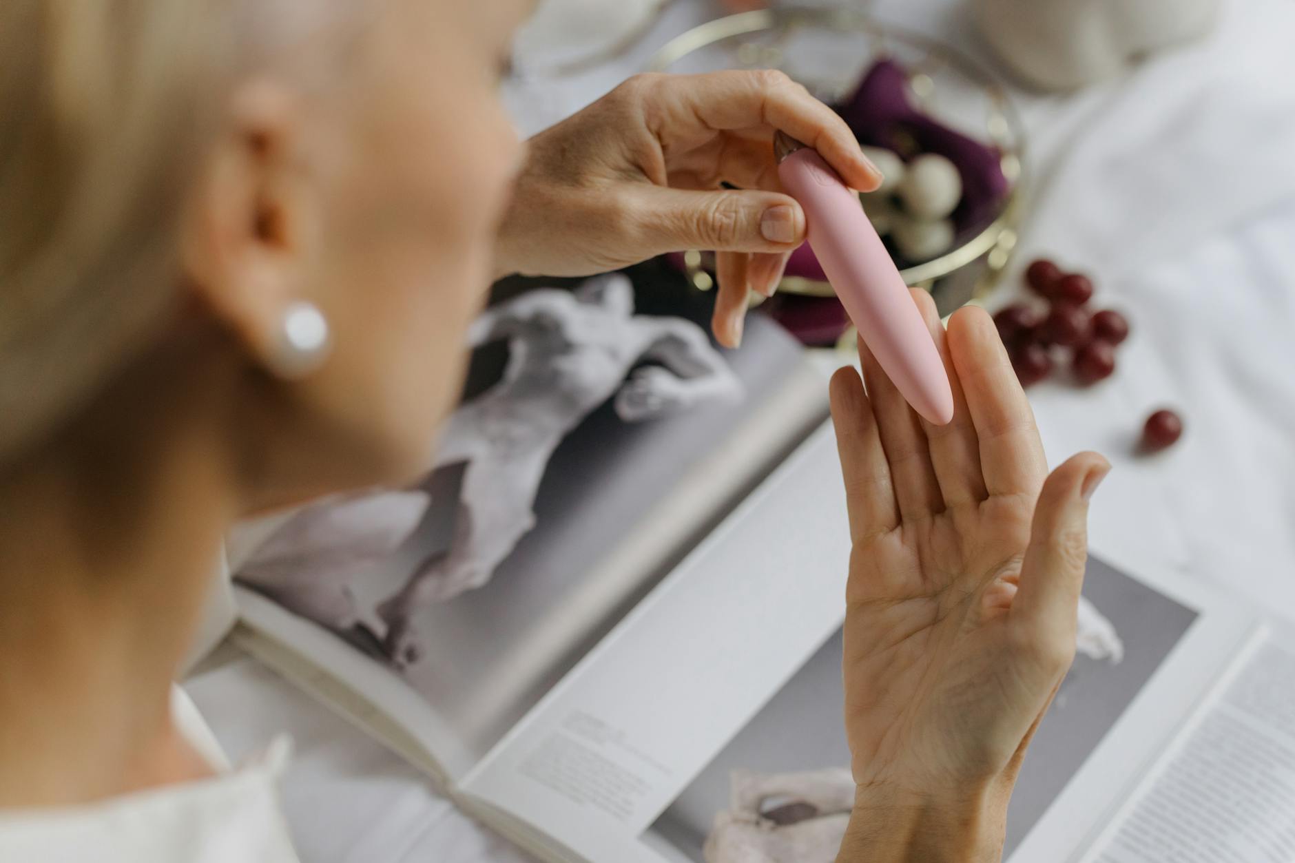 Close-up of a woman's hand holding a pink vibrator near her face, with soft lighting and art books blurred in the background.
