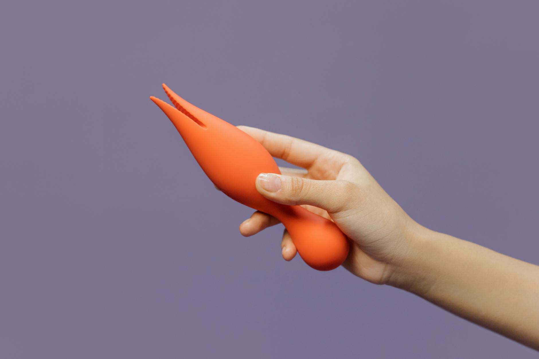 Eine Hand hält einen orangefarbenen Lemon Vibrator vor minimalistische Kulisse, die moderne Sinnlichkeit zeigt.