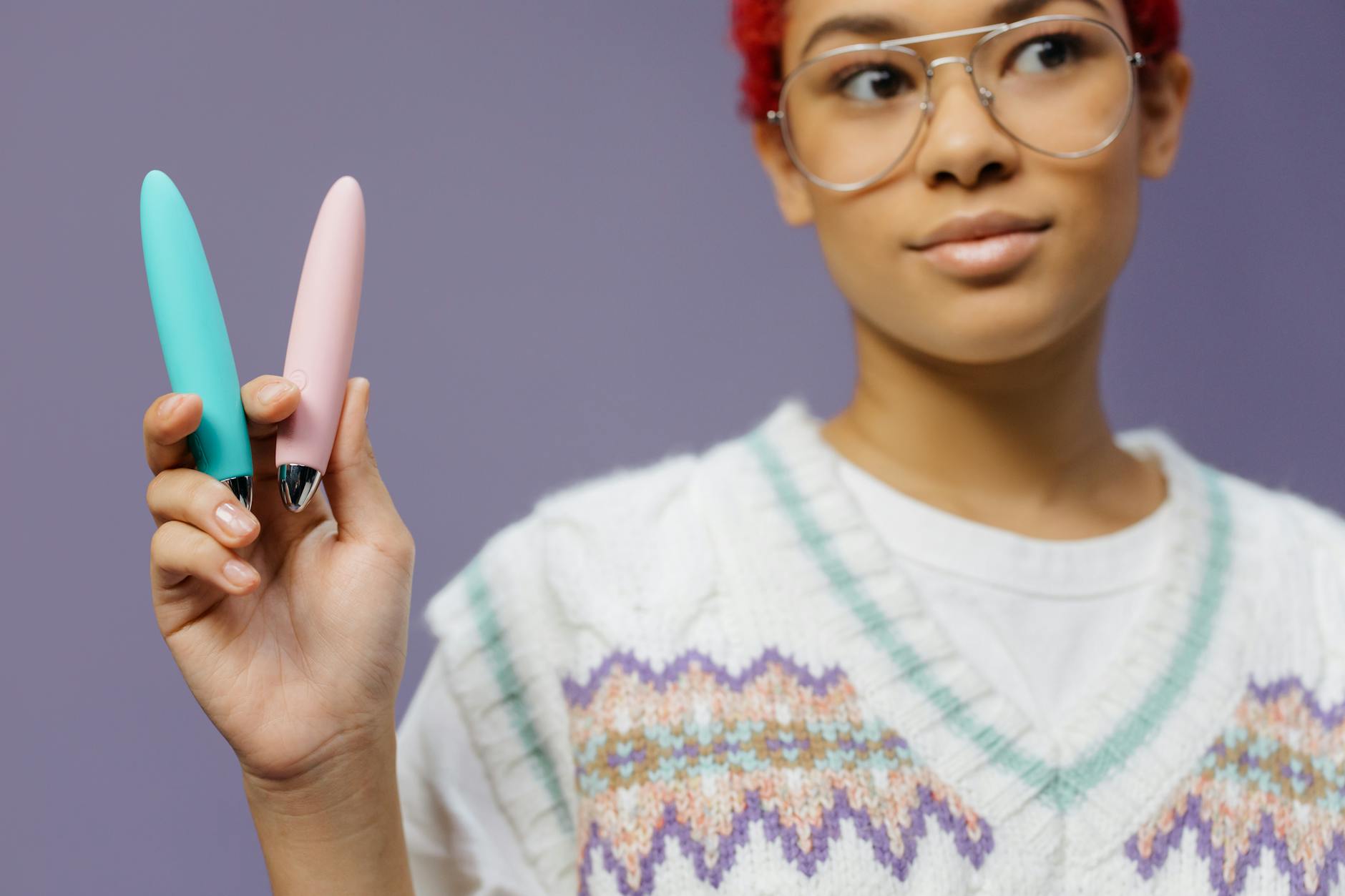 A woman in glasses holding colorful vibrators against a purple background, representing confident solo pleasure exploration.