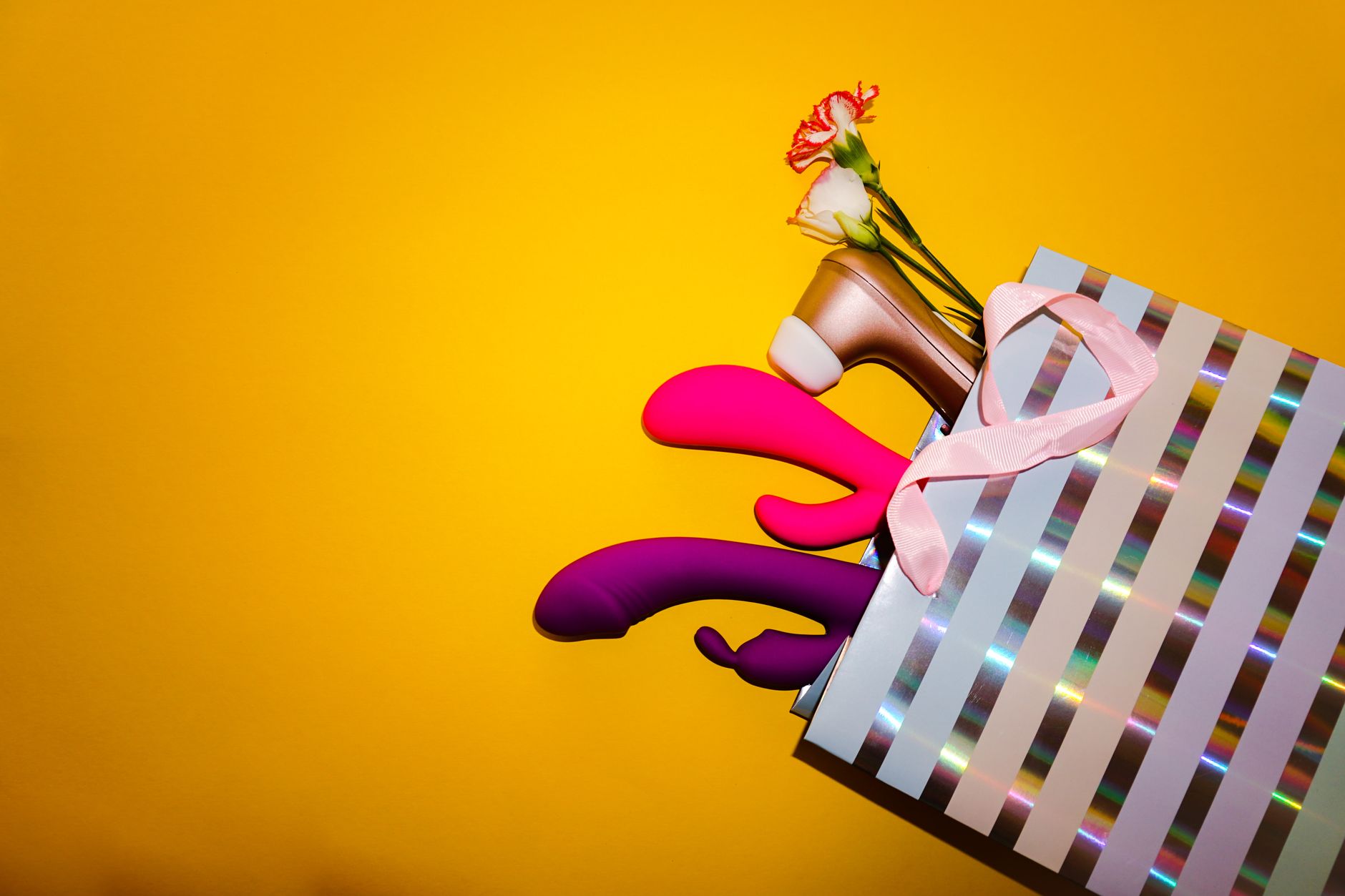 Colorful vibrators with flowers in a holographic gift bag on bold yellow background
